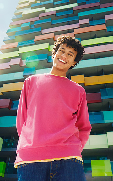 Ein junger Mann in einem rosa Pullover steht vor einem Hochhaus und lacht in die Kamera.