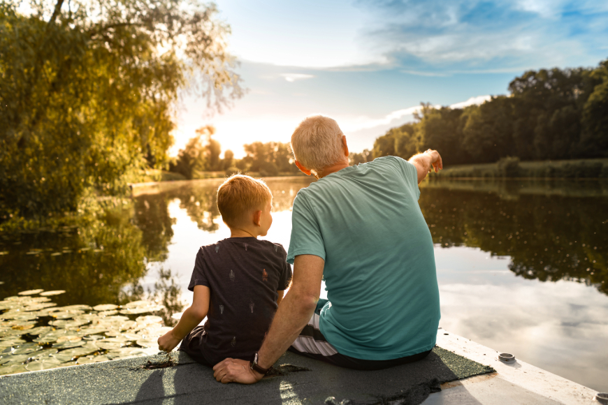 Ein älterer Mann sitzt mit einem Jungen auf einem Steg an einem See und deutet mit der rechten Hand in die Ferne.