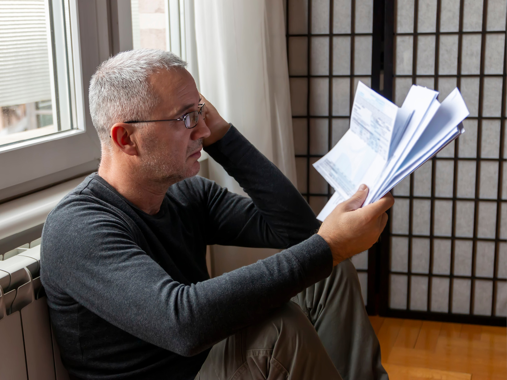 Ein Mann sitzt vor der Fensterbank am Boden, hält viele Rechnungen in der Hand und blickt darauf.