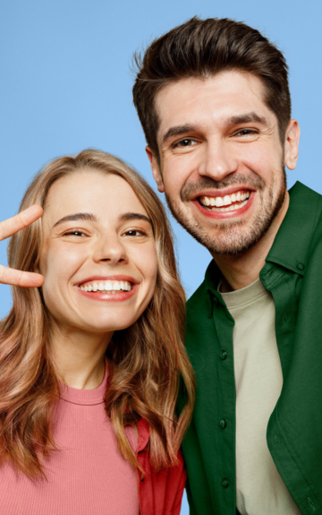 Eine junge Frau in rosa Kleidung und ein Mann in grüner Kleidung grinsen in die Kamera und zeigen ein Peace-Zeichen.