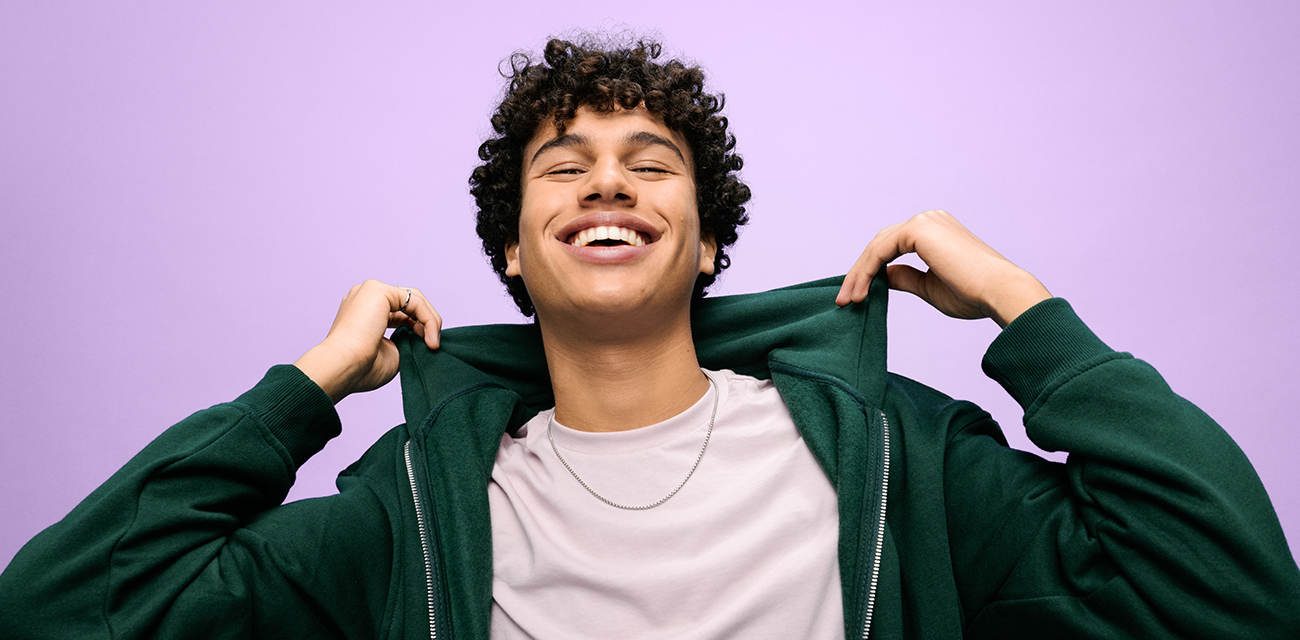 Ein junger Mann auf rosa Hintergrund richtet lachend die Kapuze seines grünen Sweatshirts.