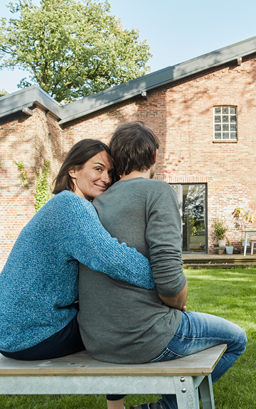 Ein Mann und eine Frau sitzen mit dem Rücken zur Kamera vor einem Haus. Die Frau umarmt den Mann von hinten. Sie blickt über die Schulter in die Kamera.