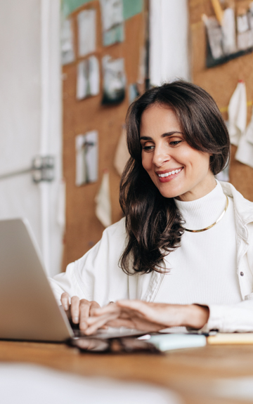 Eine lächelnde Frau im Rollkragenpullover tippt auf ihrem Laptop.