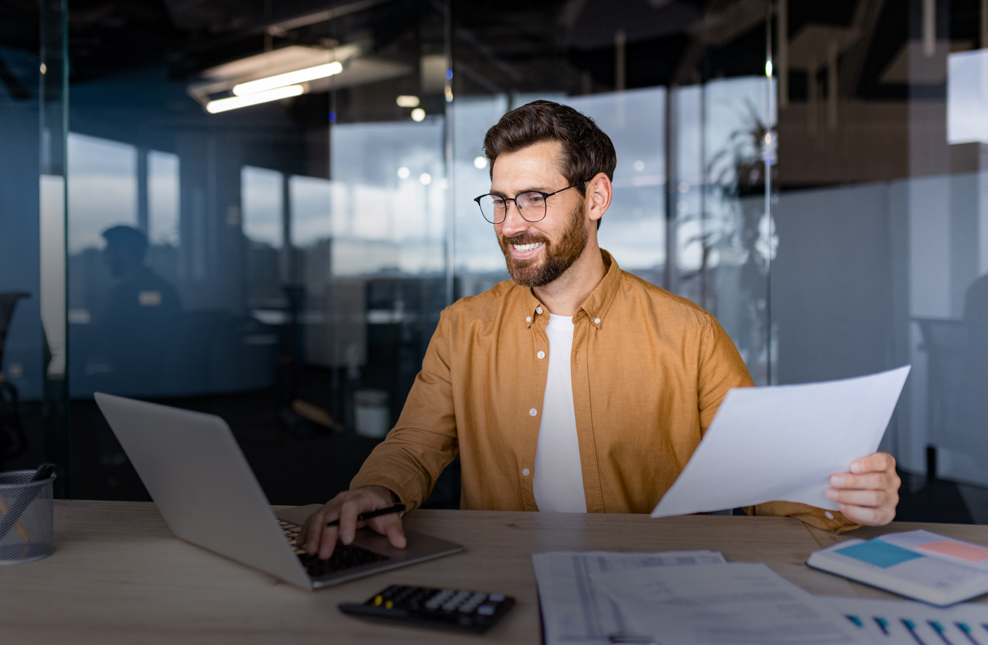Ein Mann im Büro hält ein Papier in der Hand udn blickt lächelnd auf seinen Laptop.
