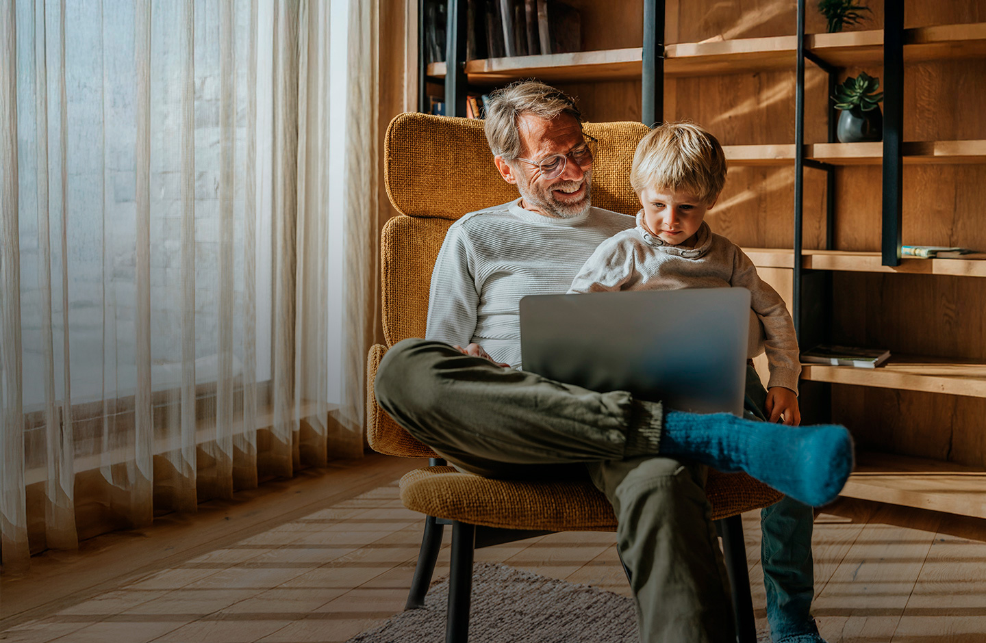 Ein Mann und ein Junge sitzen in einem Sessel und blicken gemeinsam auf ein Notebook. Der Mann lächelt, das Kind blickt konzentriert.